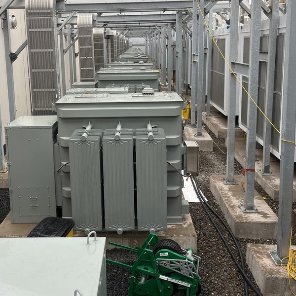 A pad-mounted transformer at the head of a long row under a steel canopy, cable runs crossing the gravel yard